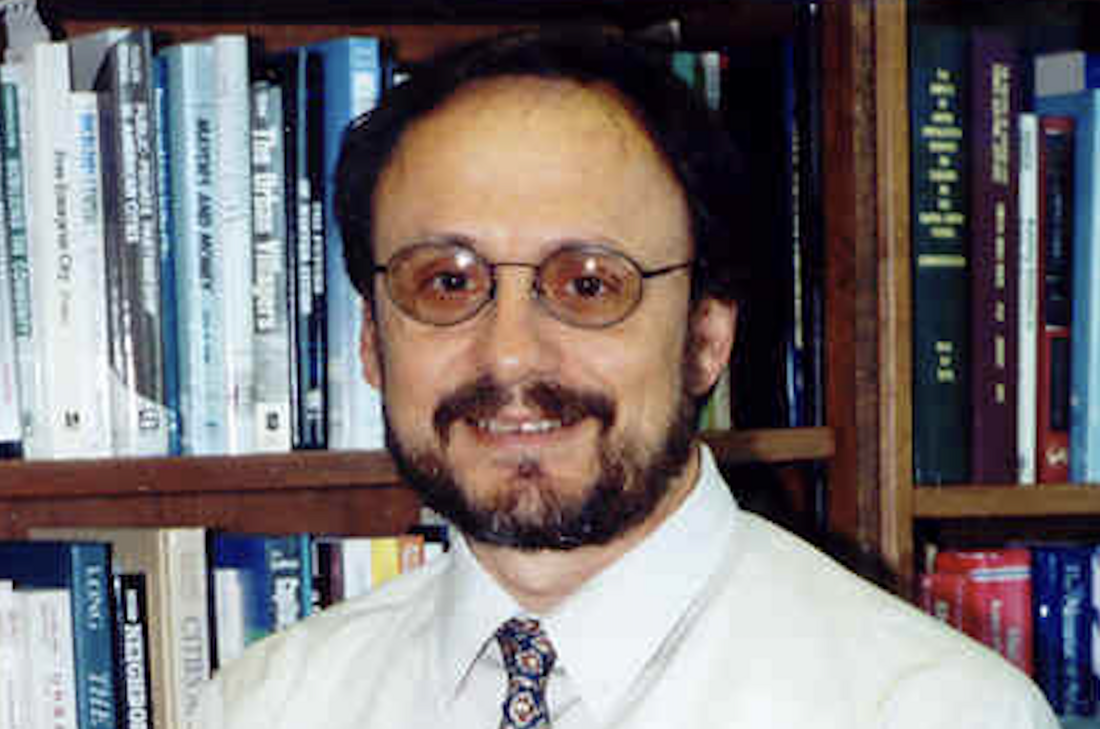 Headshot of a man with glasses and a beard smiling, with bookshelves filled with books behind him